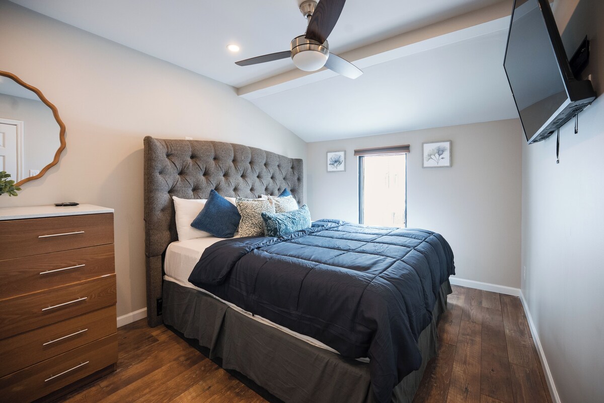 A bedroom with a tufted headboard, a bed with dark bedding, a wooden dresser, a ceiling fan, and a flat-screen TV mounted on the wall.