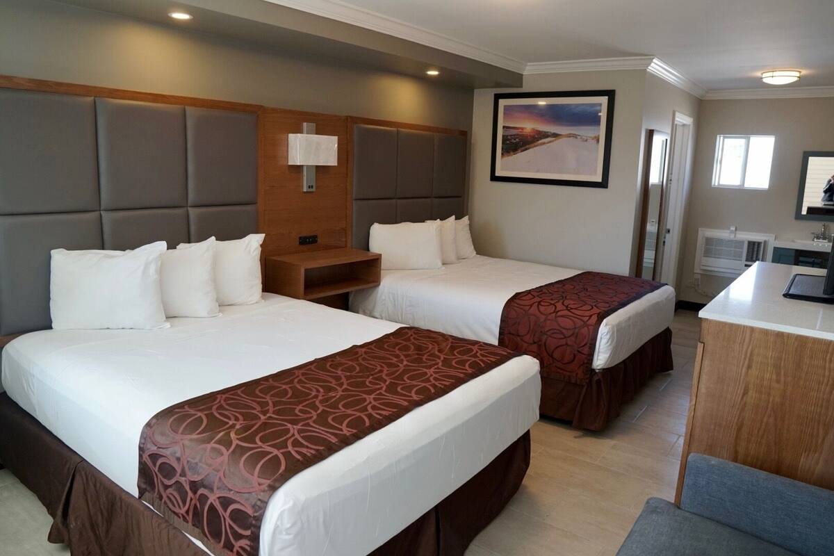 A hotel room with two beds, a wooden headboard, a bedside table, a desk, and a framed picture on the wall.