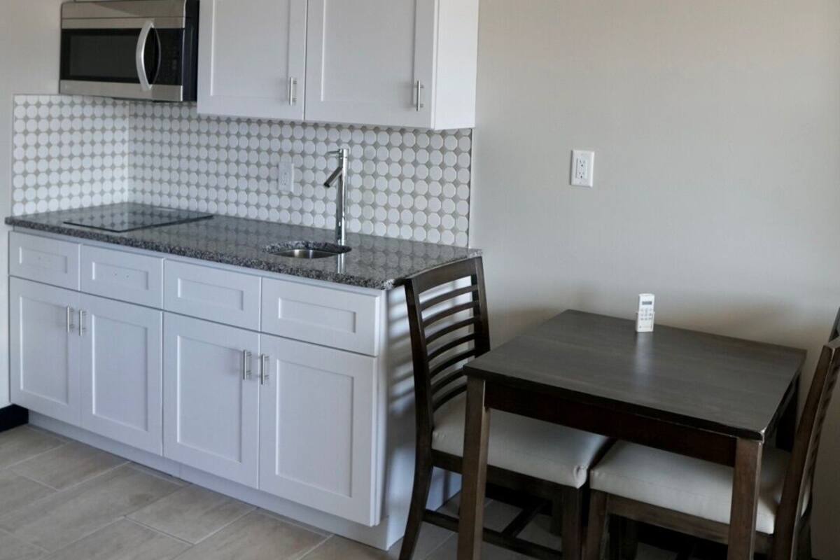 A compact kitchen with white cabinets, a microwave, and a granite countertop. There is a dining table with two chairs and a remote control on it.