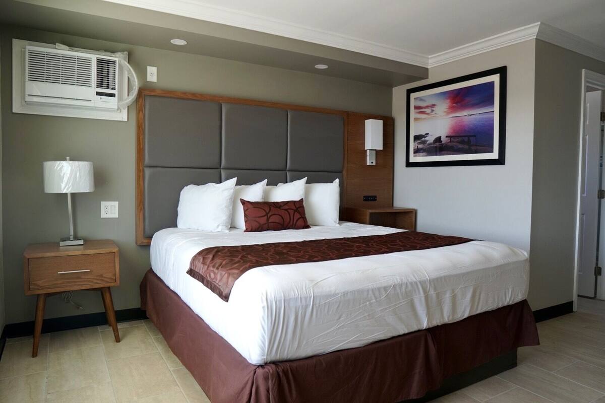 A hotel room with a large bed, a nightstand, a lamp, an air conditioning unit, a painting, and a wooden headboard.