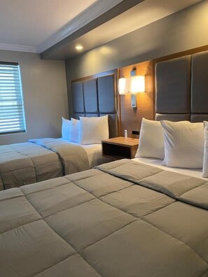 A hotel room with two beds, a nightstand, and a wall-mounted lamp. - Coastal Sands Inn (Wildwood)