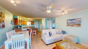A spacious living area with a white couch, dining table, and chairs. There is a kitchen with wooden cabinets and a ceiling fan. A beach-themed painting is on the wall. - Walk to Sandfest,  Shared Pools, Boardwalk to the Beach (Port Aransas)
