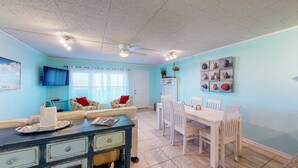 A living room with a sofa, a dining table with chairs, a TV mounted on the wall, and a ceiling fan. - Walk to Sandfest,  Shared Pools, Boardwalk to the Beach (Port Aransas)