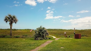 A grassy pathway leading to a beach with a palm tree and a wooden structure. - Walk to Sandfest,  Shared Pools, Boardwalk to the Beach (Port Aransas)