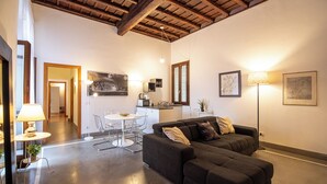 A modern living room with a dark sofa, a dining table with chairs, a kitchen area, and a ceiling with exposed beams. - Domitia Home A Pochi Passi DA Piazza DI Spagna (Roma)