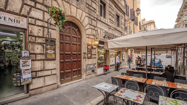 A street view with a café and outdoor seating, a shop with a large wooden door, and a building with a sign 'VENIR'. - Domitia Home A Pochi Passi DA Piazza DI Spagna (Roma)