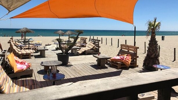 Un espace de détente aménagé sur la plage, avec des meubles en bois, des parasols et une vue sur l’océan.