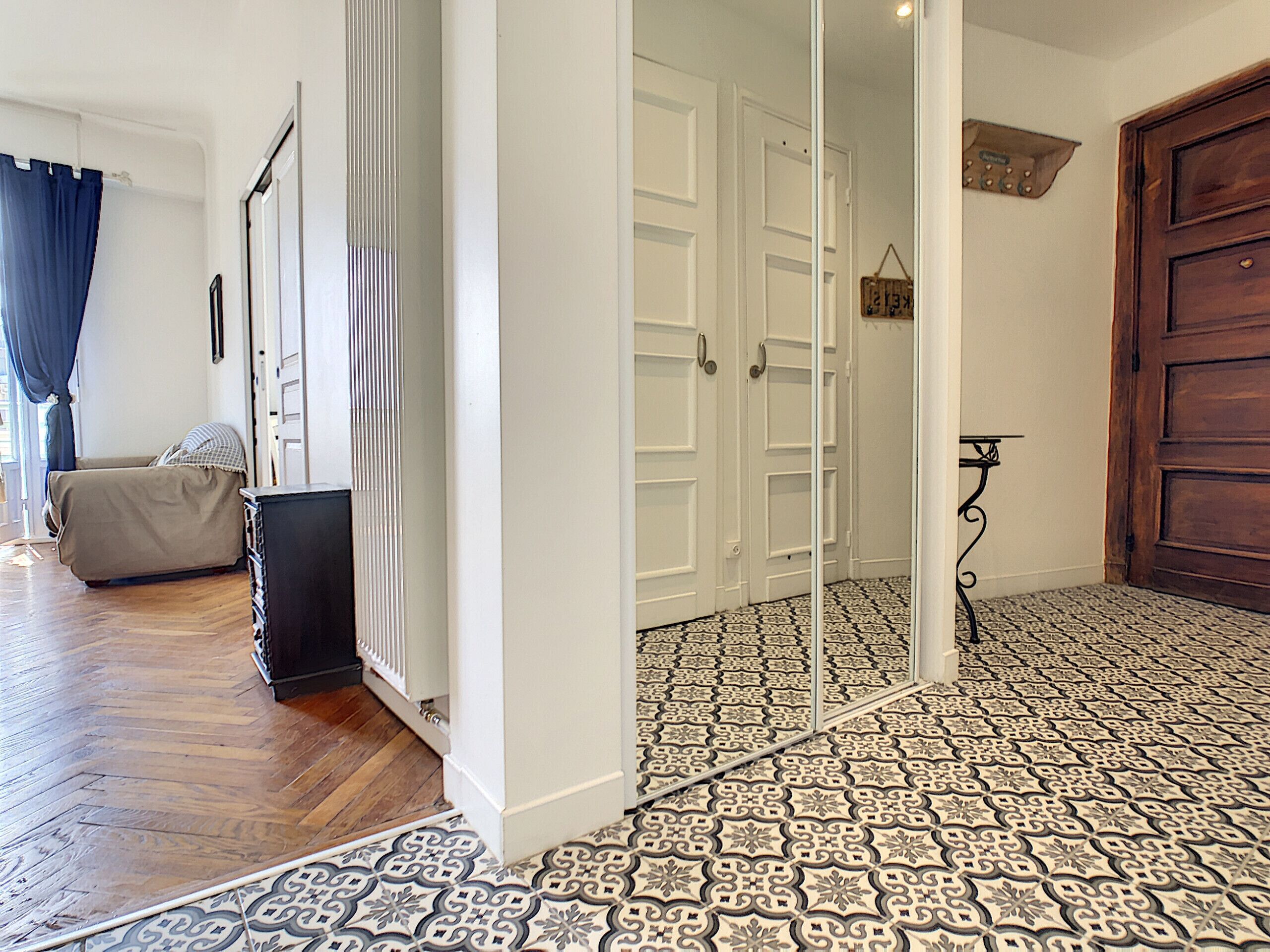 A hallway with patterned floor tiles, a wooden door, and a beige sofa in the background.