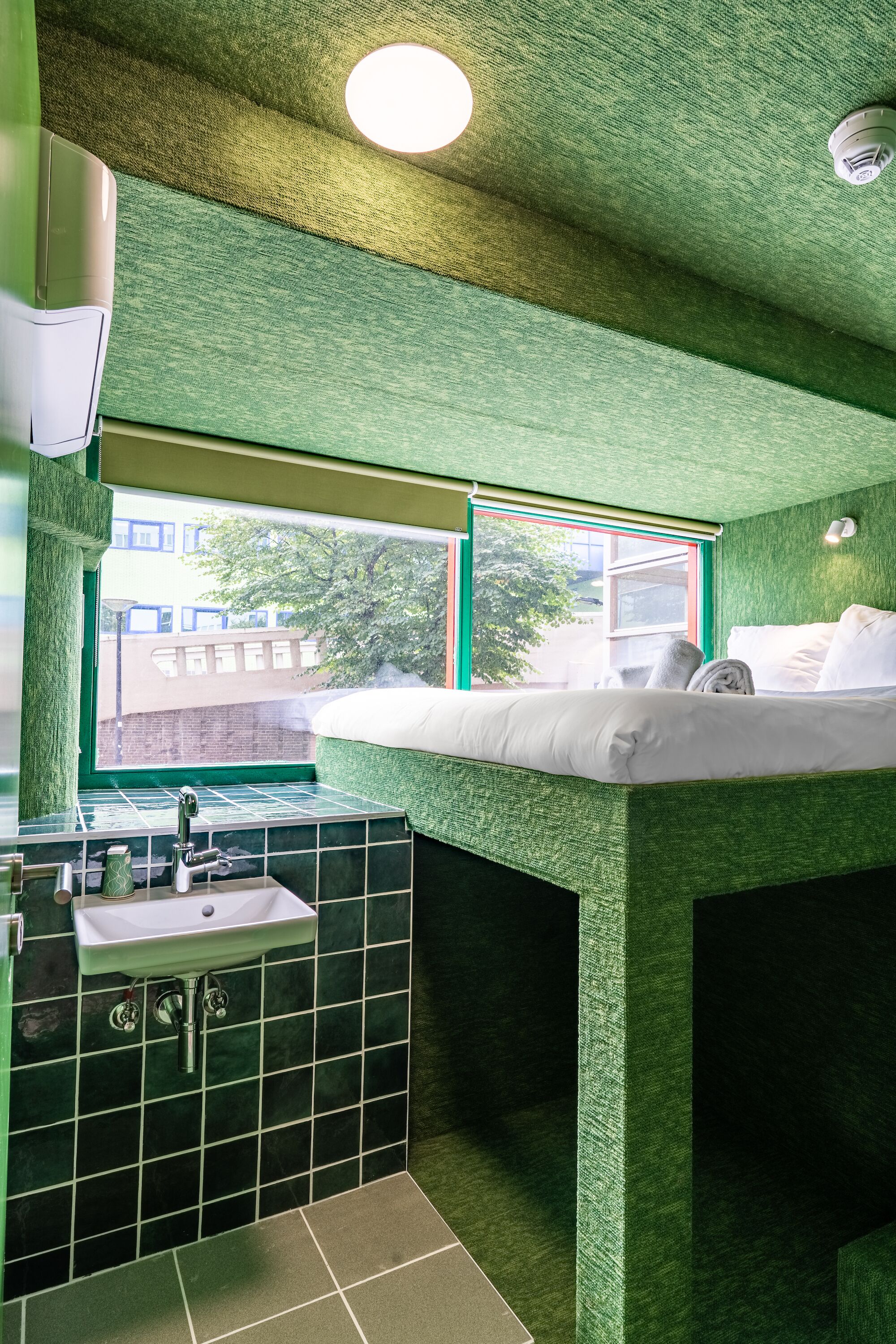 Una habitación con cama, ventana, lavamanos y pared verde con textura.