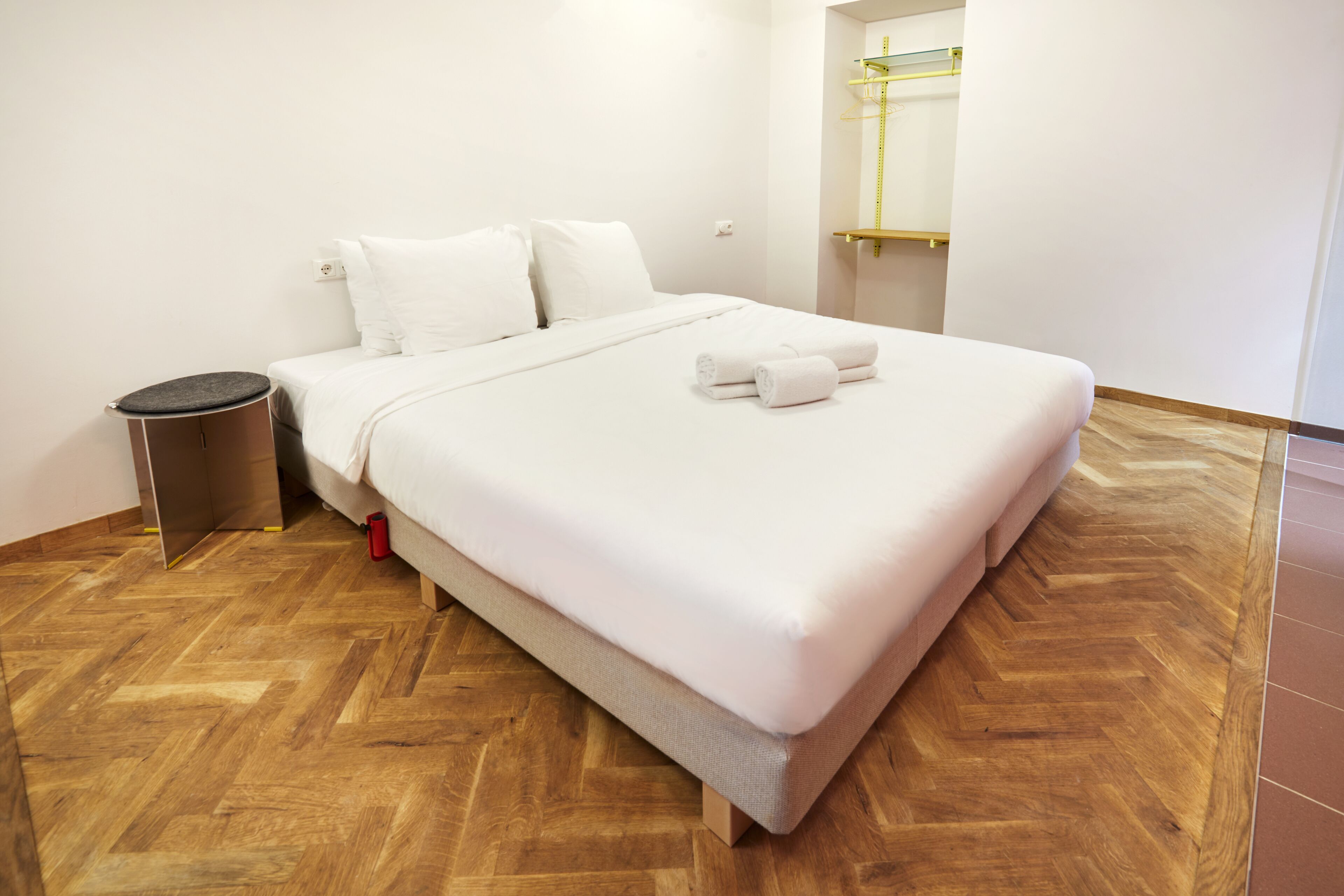 Una cama bien hecha con sábanas blancas y dos almohadas, un taburete redondo y un cesto de basura.