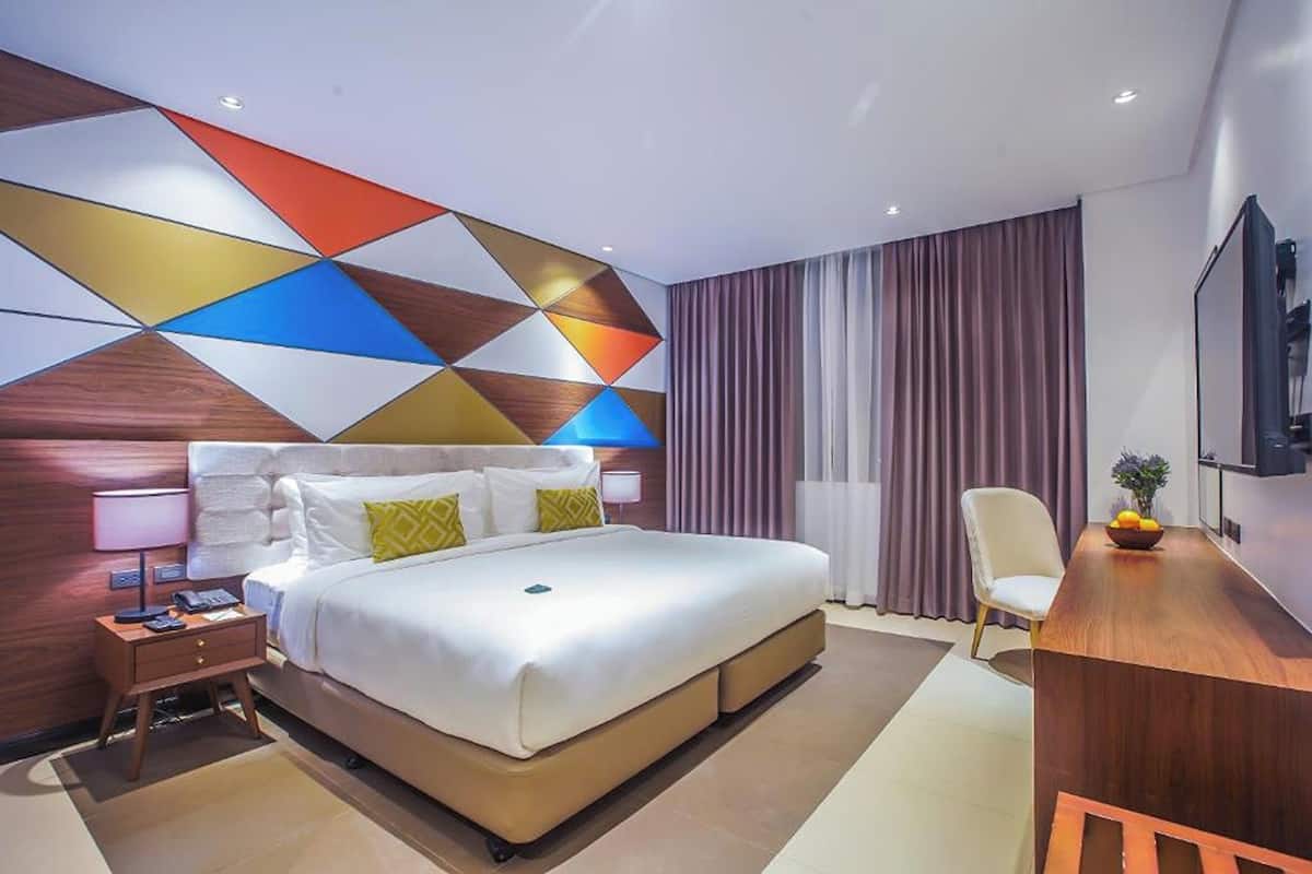 A hotel room with a large bed, a desk, a chair, a TV, and a wall with a geometric pattern.
