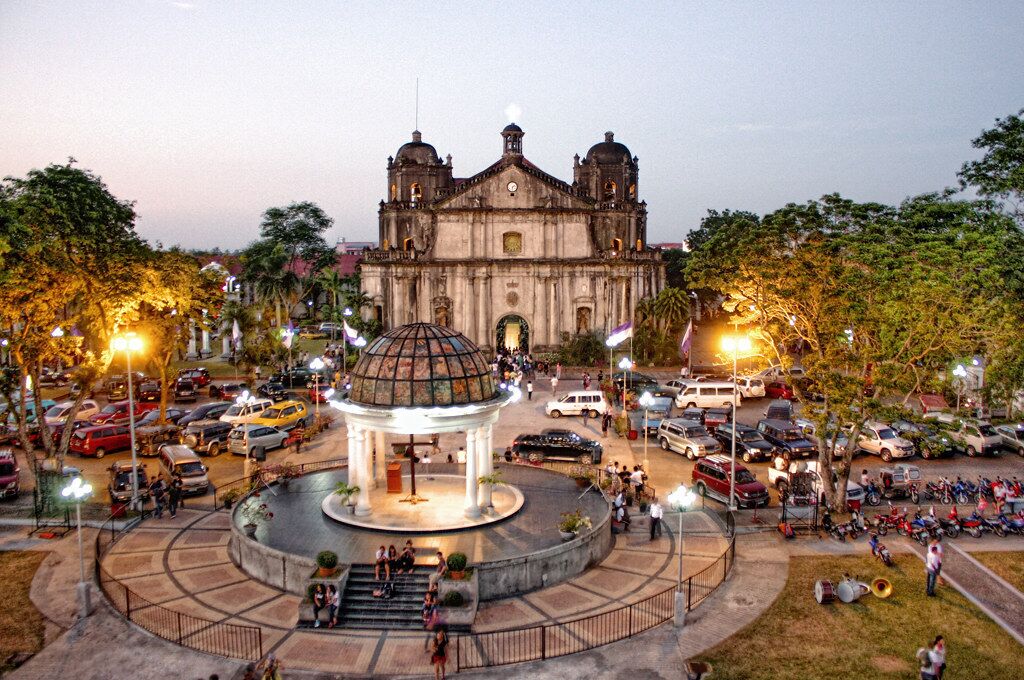 Um edifício histórico com uma cúpula central e duas torres sineiras, rodeado por uma praça circular com uma fonte e áreas de estar.