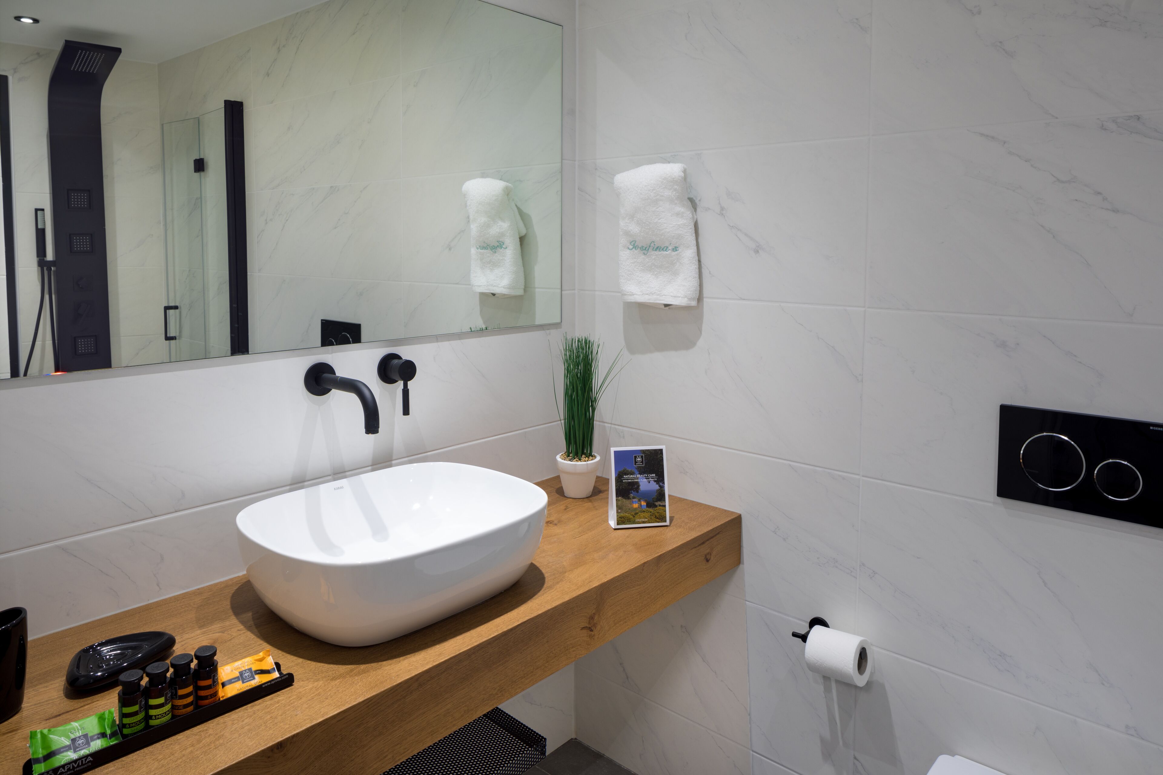A modern bathroom with a large white sink, a wooden countertop, a mirror, a towel rack, a potted plant, and a toilet paper holder.