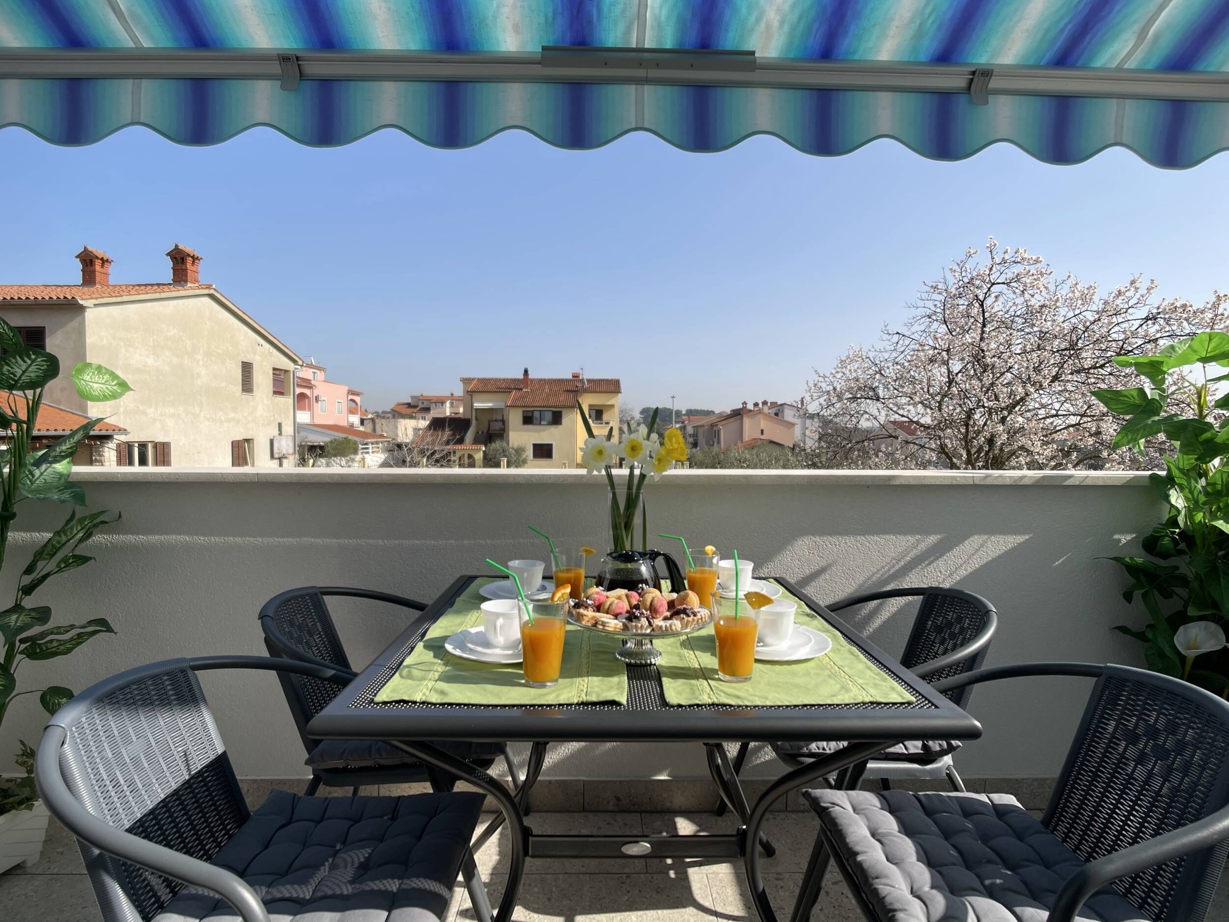 A terrace with a table set for breakfast, overlooking a residential area with houses and flowering trees.