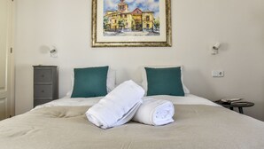 Ein ordentlich bezogenes Bett mit zwei gerollten weißen Handtüchern, ein gerahmtes Bild eines Gebäudes an der Wand und ein kleiner Nachttisch mit einem Buch.