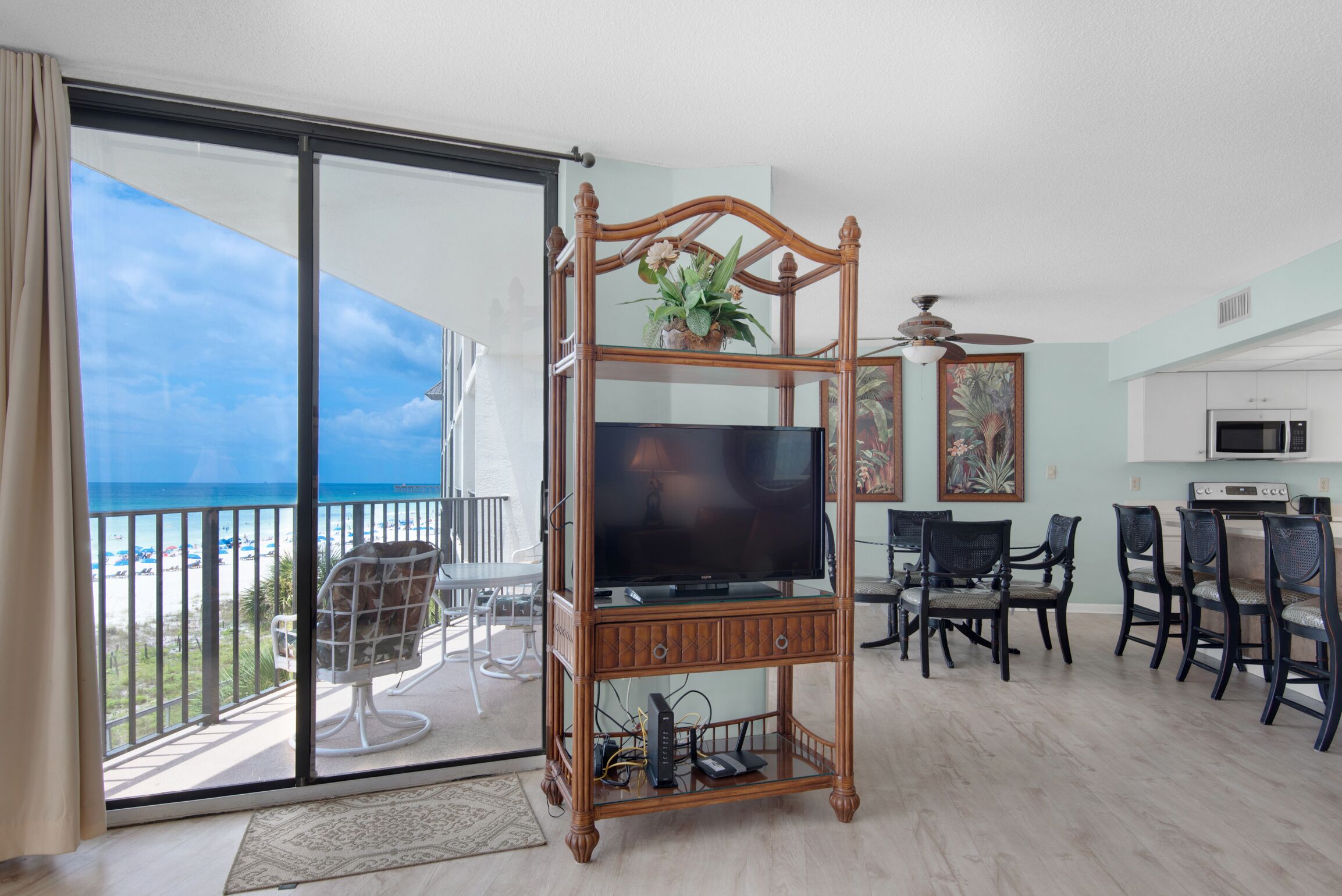 A spacious living area with a large TV on a wooden stand, a dining table with chairs, a kitchen area, and a balcony with a view of the beach.