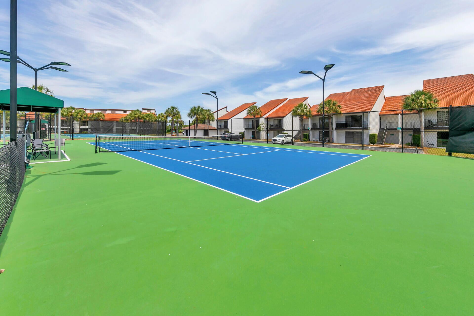 En utomhus tennisbana med en grön och blå yta, omgiven av byggnader med röda tak och en klar himmel.
