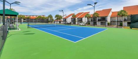 En utomhus tennisbana med en grön och blå yta, omgiven av byggnader med röda tak och en klar himmel.