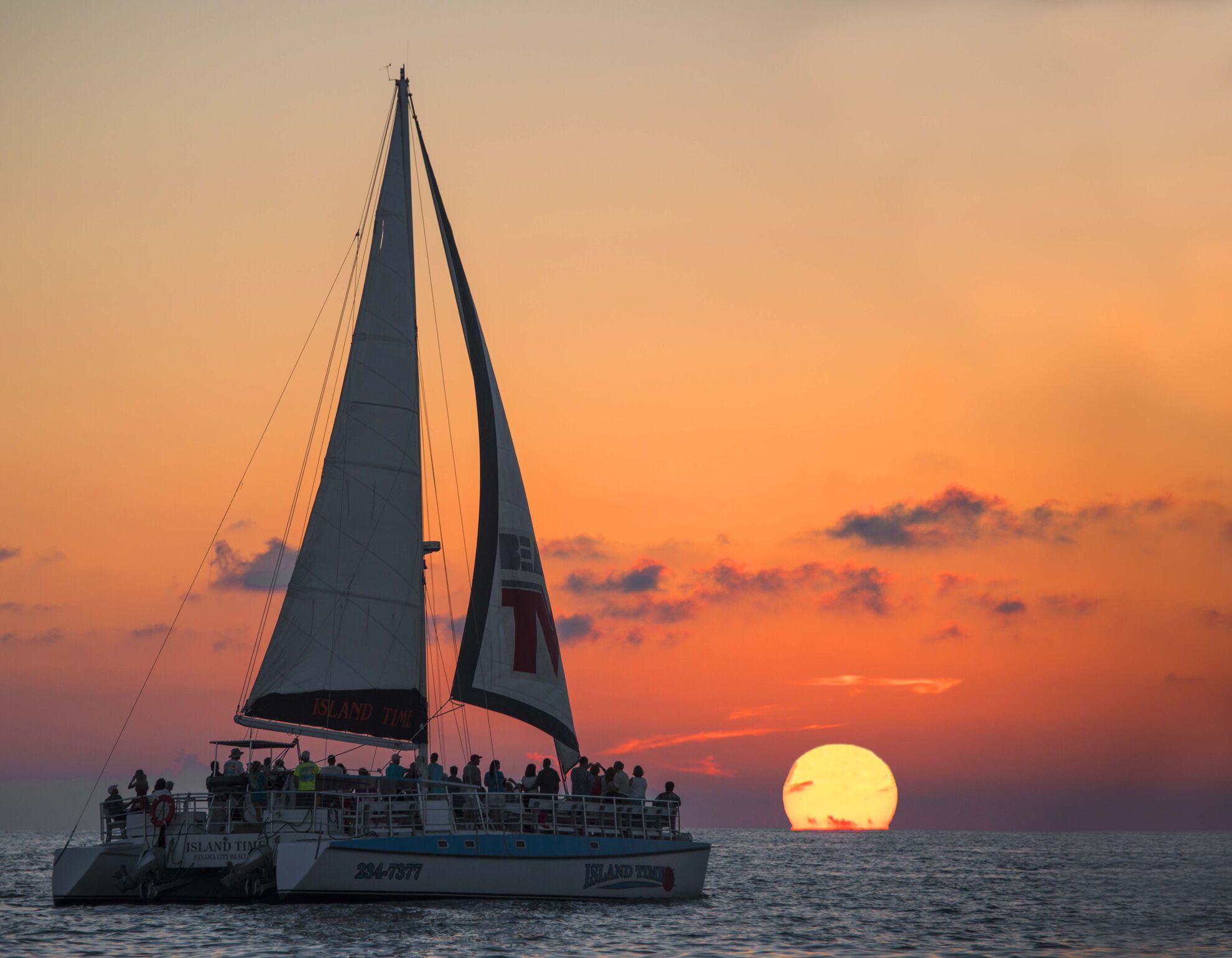 A sailboat with passengers, sailing at sunset with the sun visible in the background.