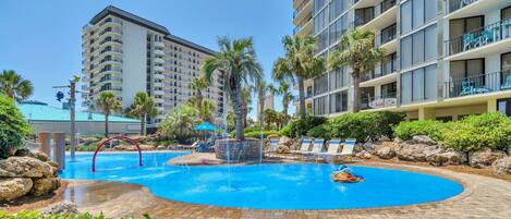 A swimming pool area with a water feature, palm trees, and a multi-story hotel building in the background.