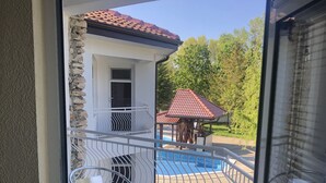 A balcony with a metal table and chairs, a stone wall, and a view of a swimming pool and trees. - Bultrak- eco village (Balchik)