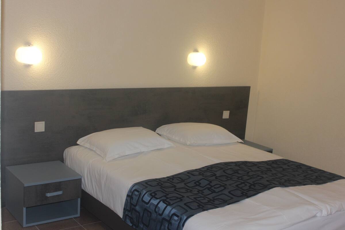 Uma cama bem arrumada com um edredom estampado, uma mesinha de cabeceira com gaveta e duas luminárias fixadas na parede.