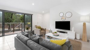 Un salon moderne avec un canapé gris, un téléviseur à écran plat et un balcon offrant une vue sur les arbres.