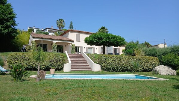 A two-story house with a swimming pool, surrounded by well-maintained hedges and a large rock.