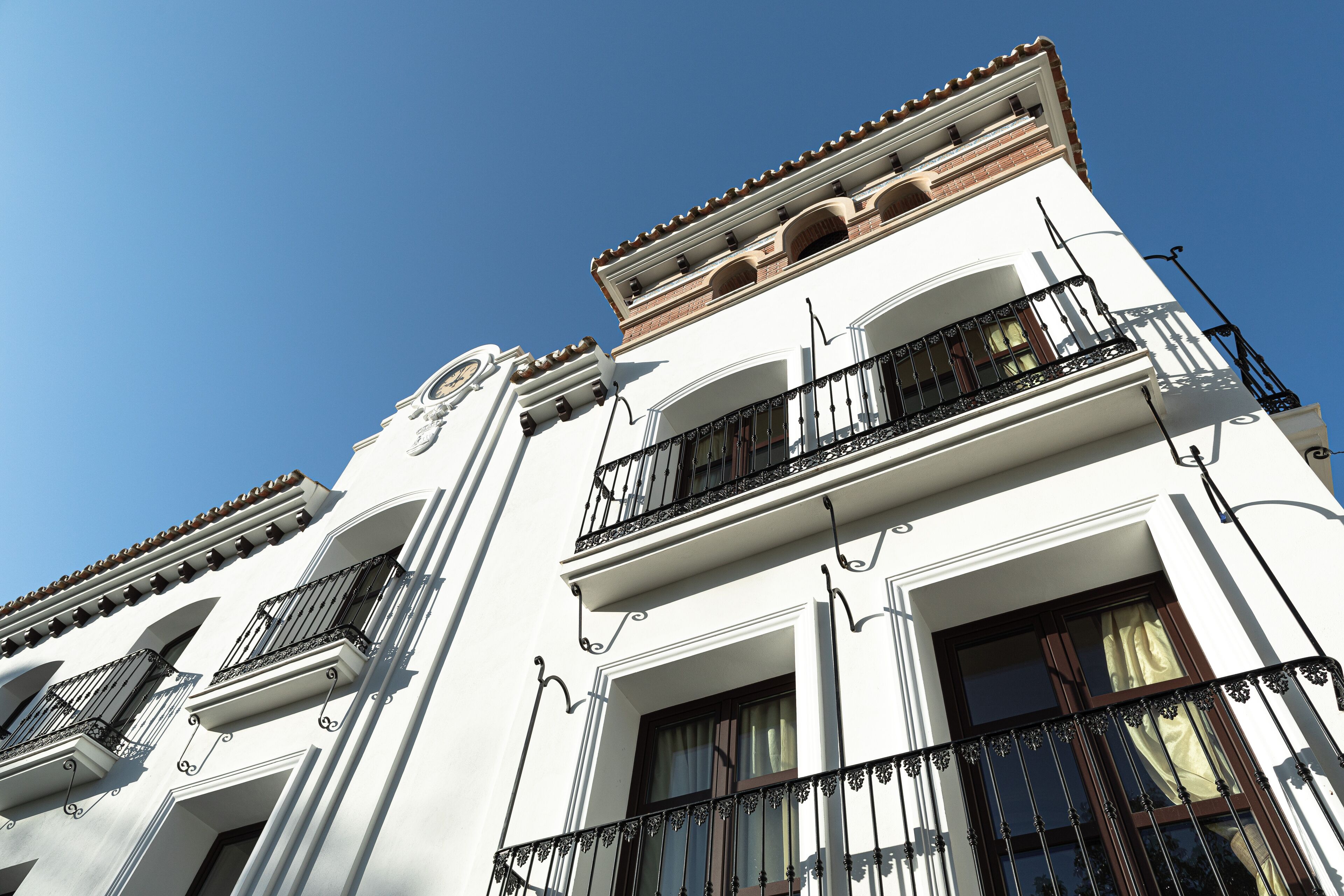 Un edificio de varios pisos con fachadas blancas, múltiples balcones con barandillas de hierro forjado y un cielo azul despejado al fondo.