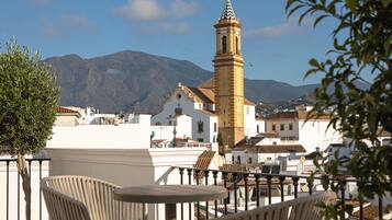A terrace with chairs and a table overlooking a town with a prominent tower and mountains in the background.