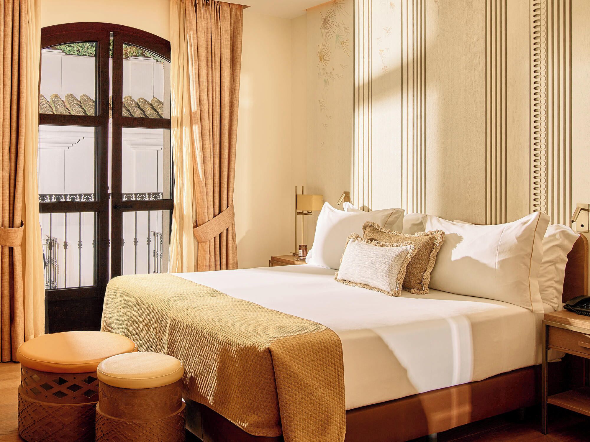 A hotel room with a bed, bedside table, lamp, and two ottomans.