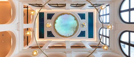 Ein großzügiges Atrium mit einem zentralen, runden Oberlicht, mehreren Balkonen und hängenden, kugelförmigen Leuchten.