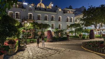 Un edificio histórico con cúpula y arquitectura clásica, rodeado de un jardín cuidado con senderos de piedra y plantas en macetas.