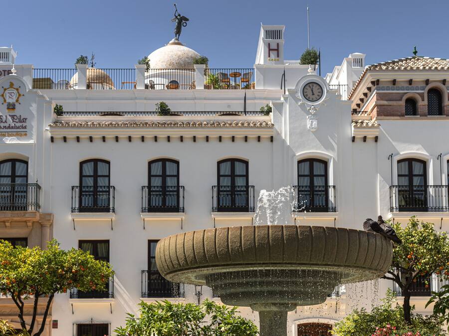 A white building with a fountain in front, balconies, and a clock.
