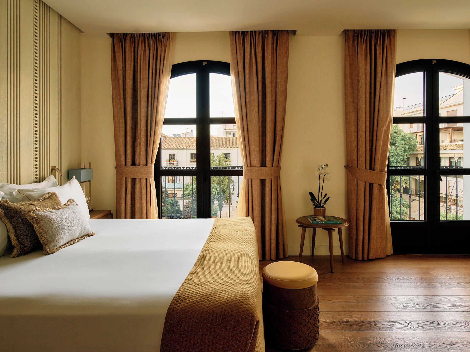 A hotel room with a large bed, a small table with a plant, and large windows with curtains.