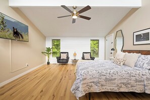 A bedroom with a bed, two chairs, a ceiling fan, and a large window.