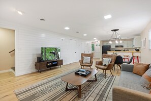A modern living room with a flat-screen TV, leather chairs, a coffee table, and a kitchen in the background.