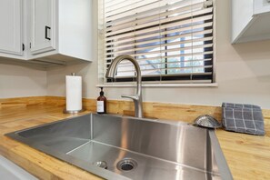 Un lavamanos de acero inoxidable con una llave moderna, una encimera de madera, una ventana con persianas y un perchero para toallas.