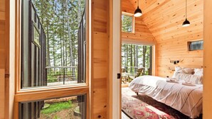 A wooden cabin interior with a bed, a window overlooking a forest, and a balcony.