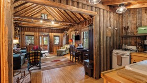 A rustic cabin interior with a wooden floor, a dining area, a kitchen with a stove, and a cozy seating area with a fireplace.