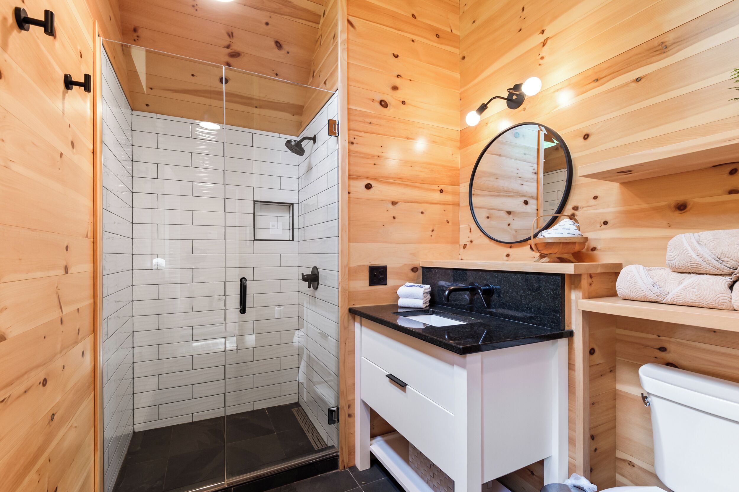 A wooden cabin bathroom with a glass-enclosed shower, a white sink with a black countertop, and a white toilet.