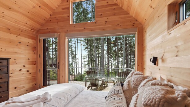 A wooden cabin bedroom with a large bed, a cozy seating area, and a balcony with a view of tall trees.