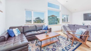 A spacious living room with a brown leather sofa, a wooden coffee table, and a patterned rug. Large windows offer a view of a residential area.