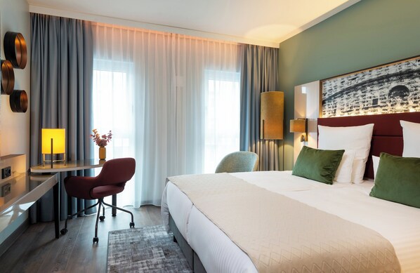 Habitación de hotel con cama, escritorio, silla y ventanas amplias con cortinas.