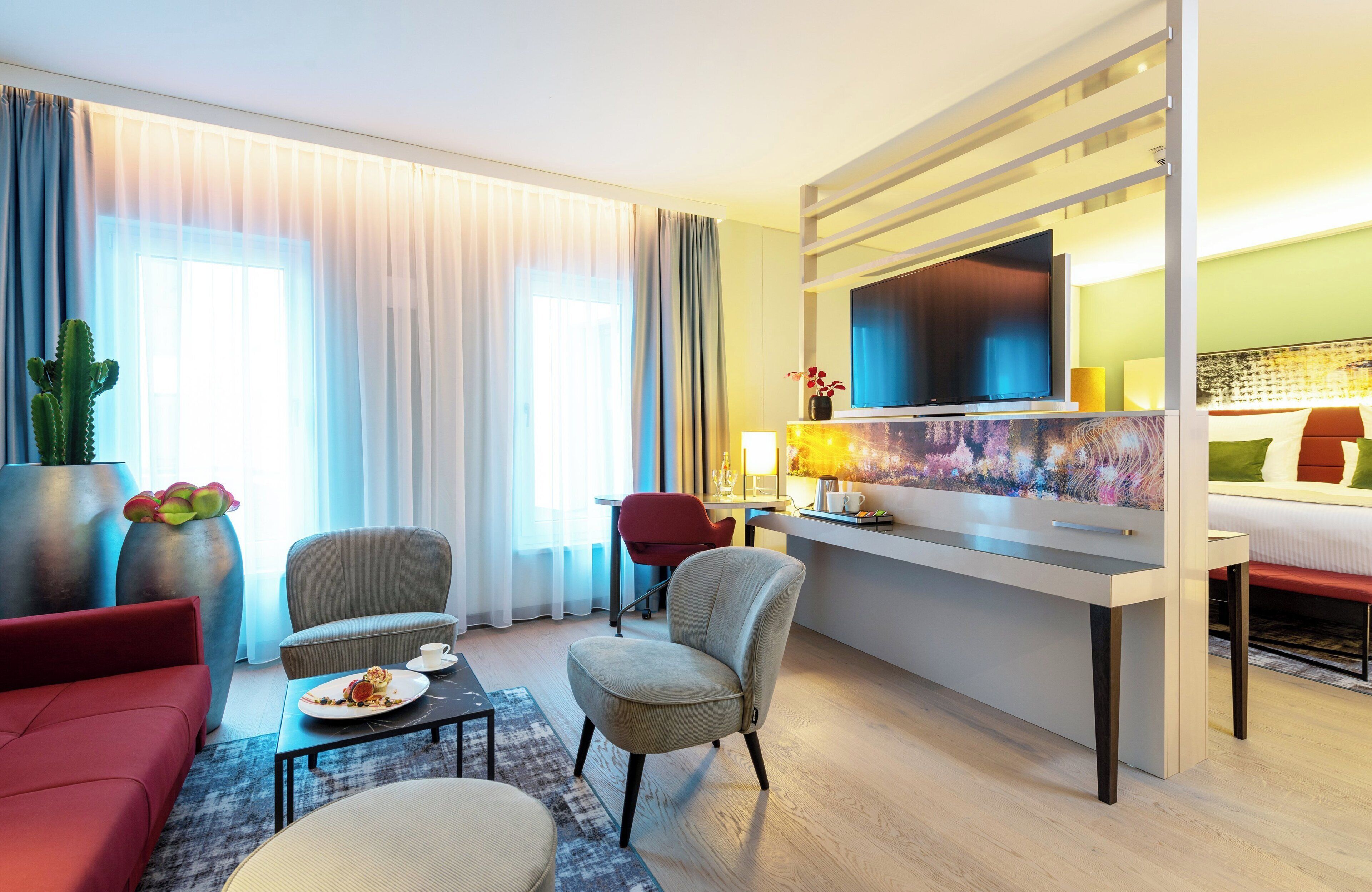 Una habitación de hotel moderna con televisión de pantalla plana, un área de comedor con mesa y sillas, un sofá y un amplio ventanal con cortinas.