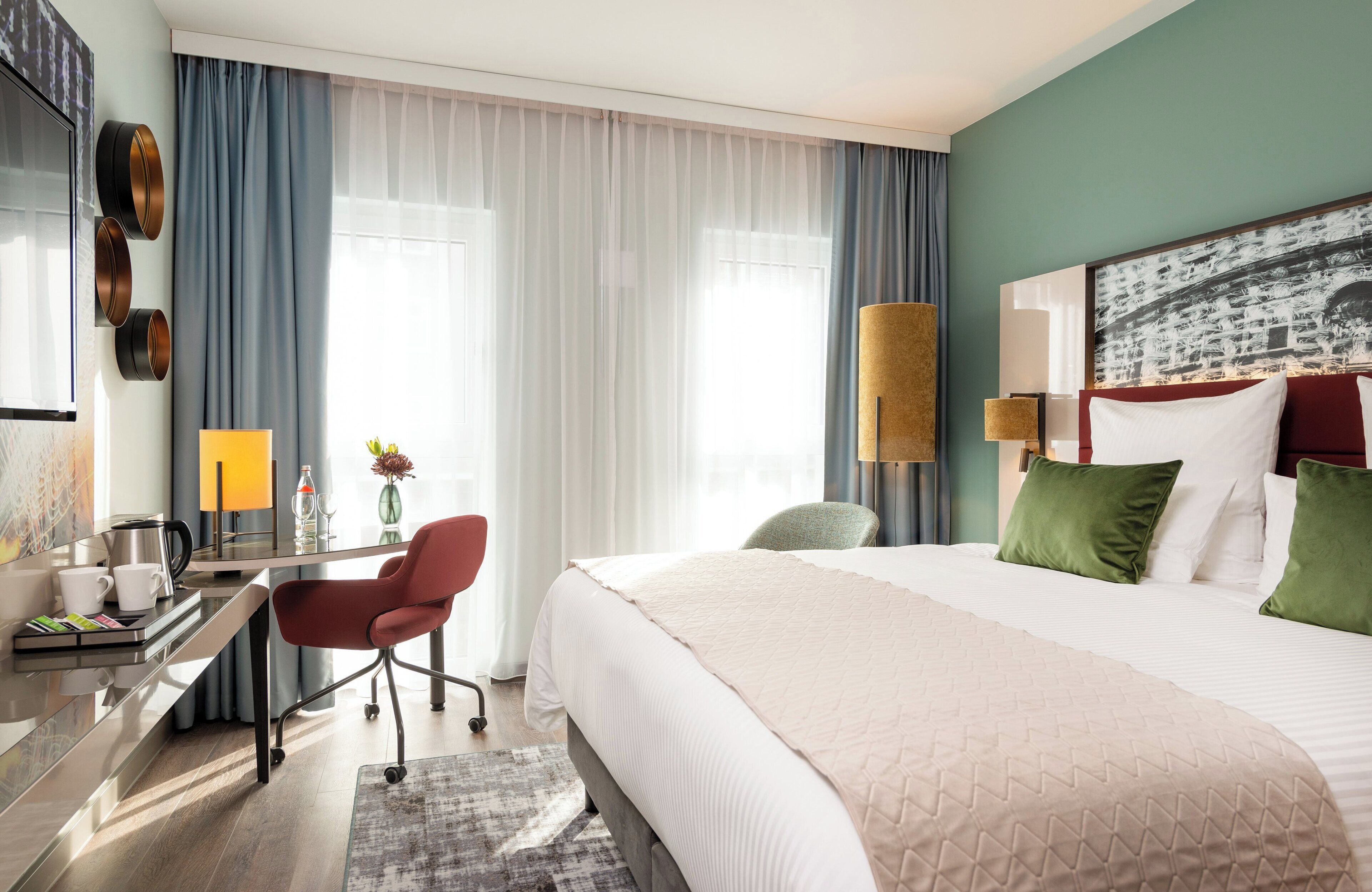 A hotel room with a bed, desk, chair, and a painting on the wall.