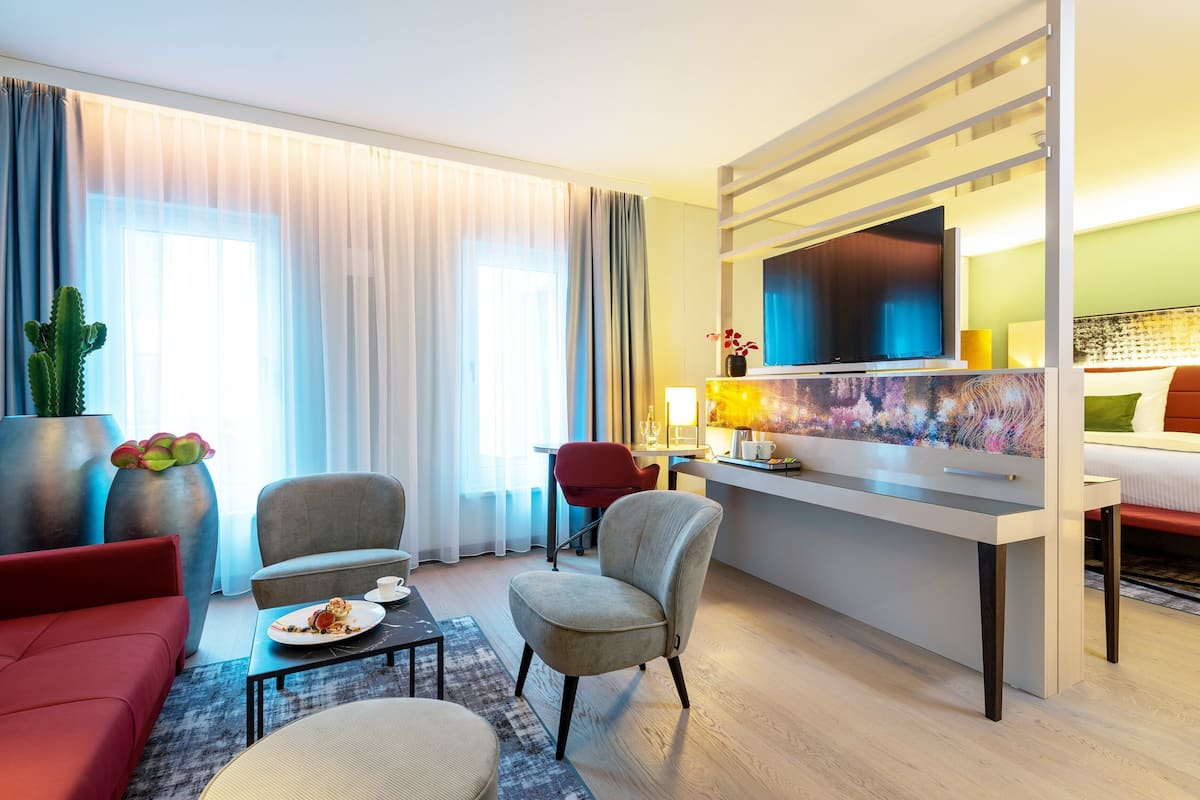 A modern hotel room with a large window, a flat-screen TV, a desk, and a comfortable seating area.