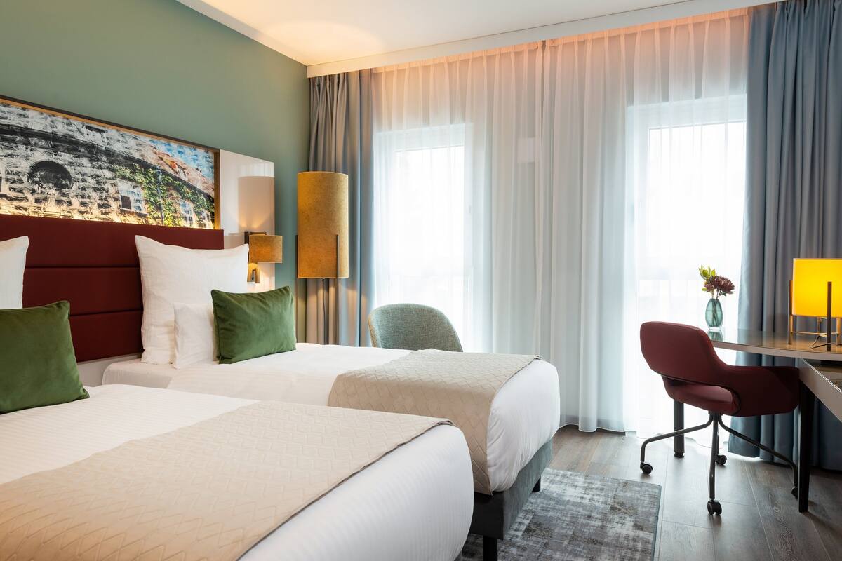 A hotel room with two beds, a desk, a chair, a lamp, and a painting on the wall.