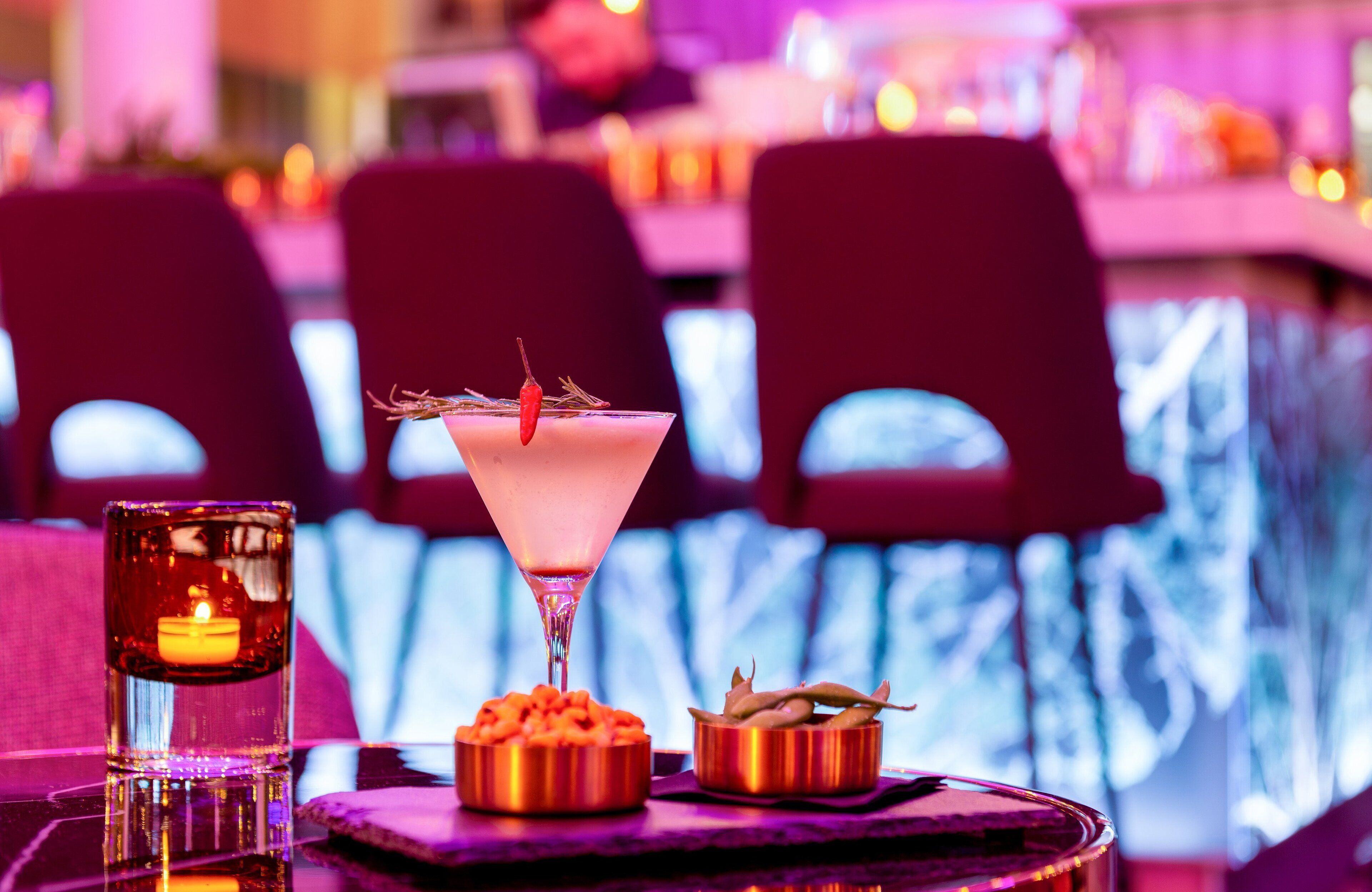 A bar setting with a cocktail, a candle, and two bowls of snacks.