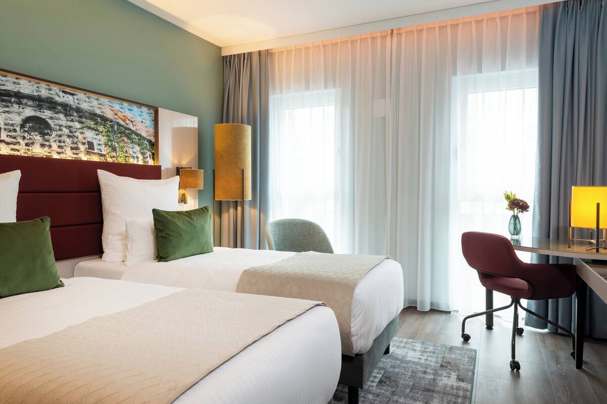A hotel room with two beds, a desk, a chair, a lamp, and a painting on the wall.